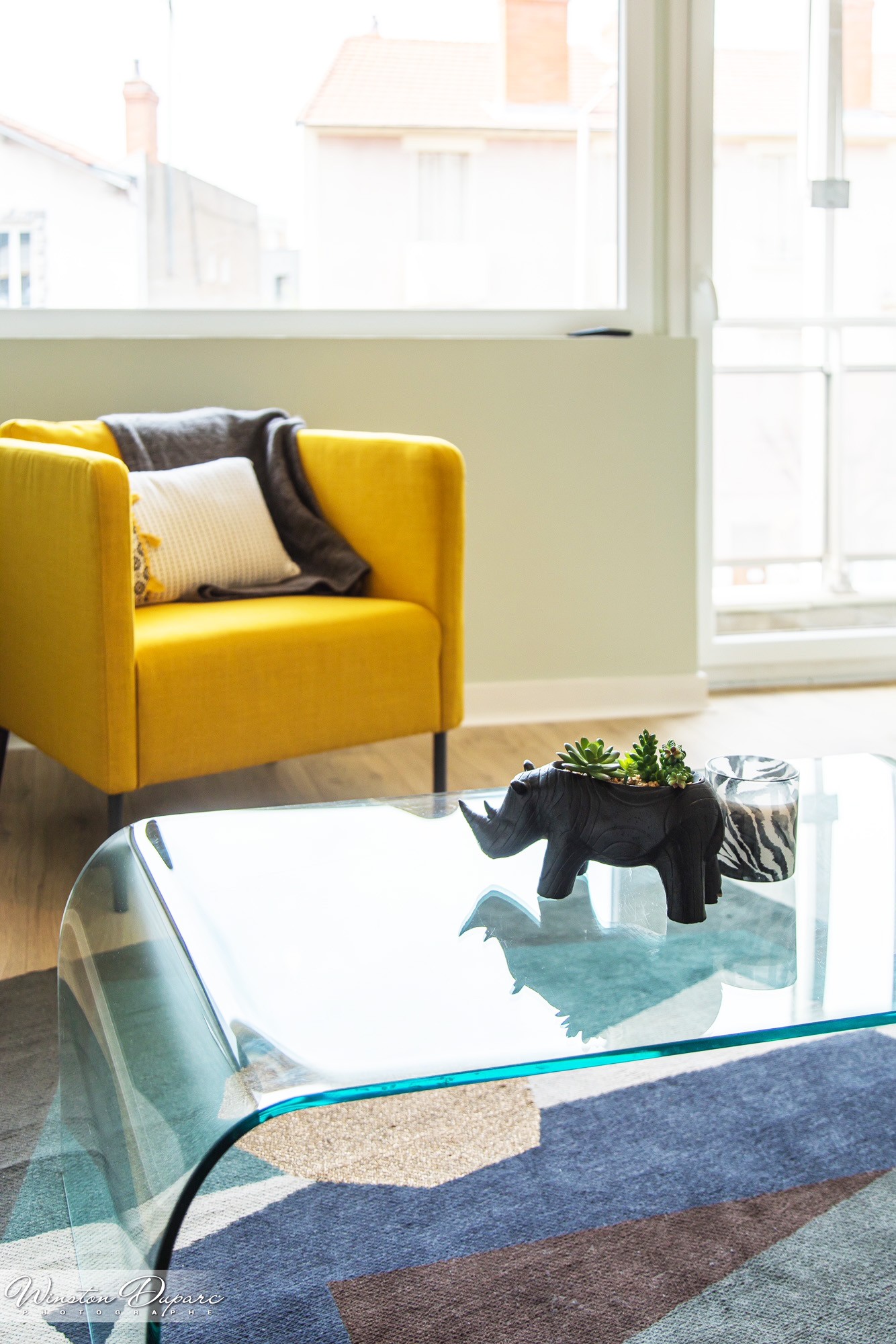 Photo d'une partie du salon montrant un fauteuil jaune avec des coussin et une table basse en verre