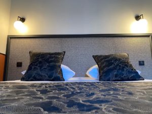 Photo en face du lit qui permet de voir les coussins du lit avec la tête de lit derrière et les interrupteur de chaque coté ainsi que les luminaires sur le mur en haut
